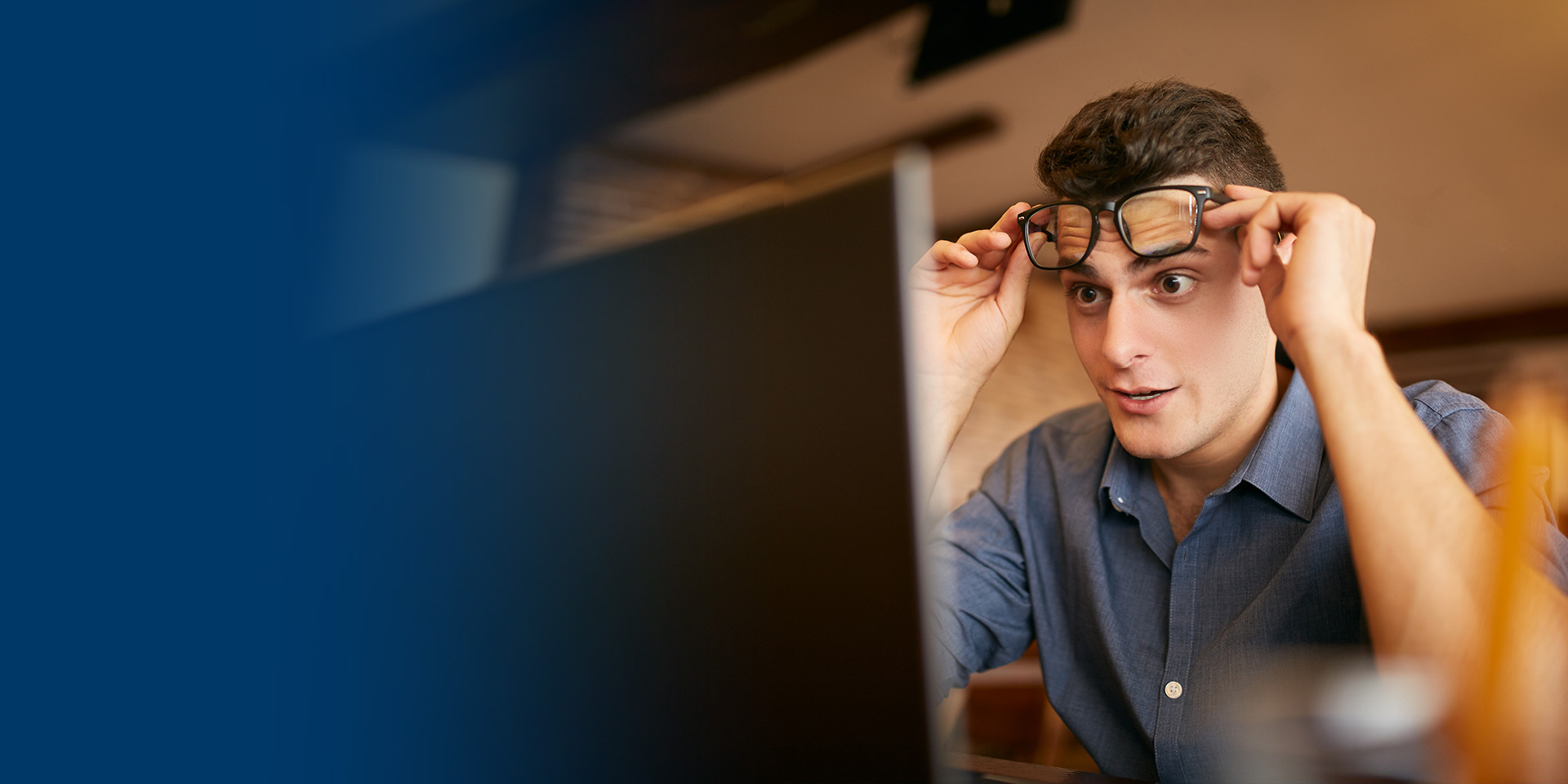 A young man sits at his computer and raises his glasses in surprise for what is on the screen