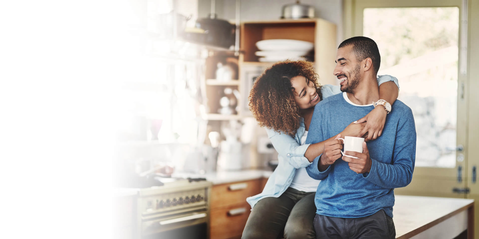 A couple hugging in their kitchen while he drinks coffee